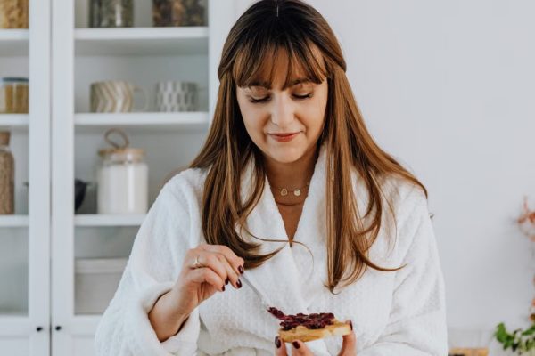 Woman in a bathrobe spreading jam on toast in a bright kitchen.